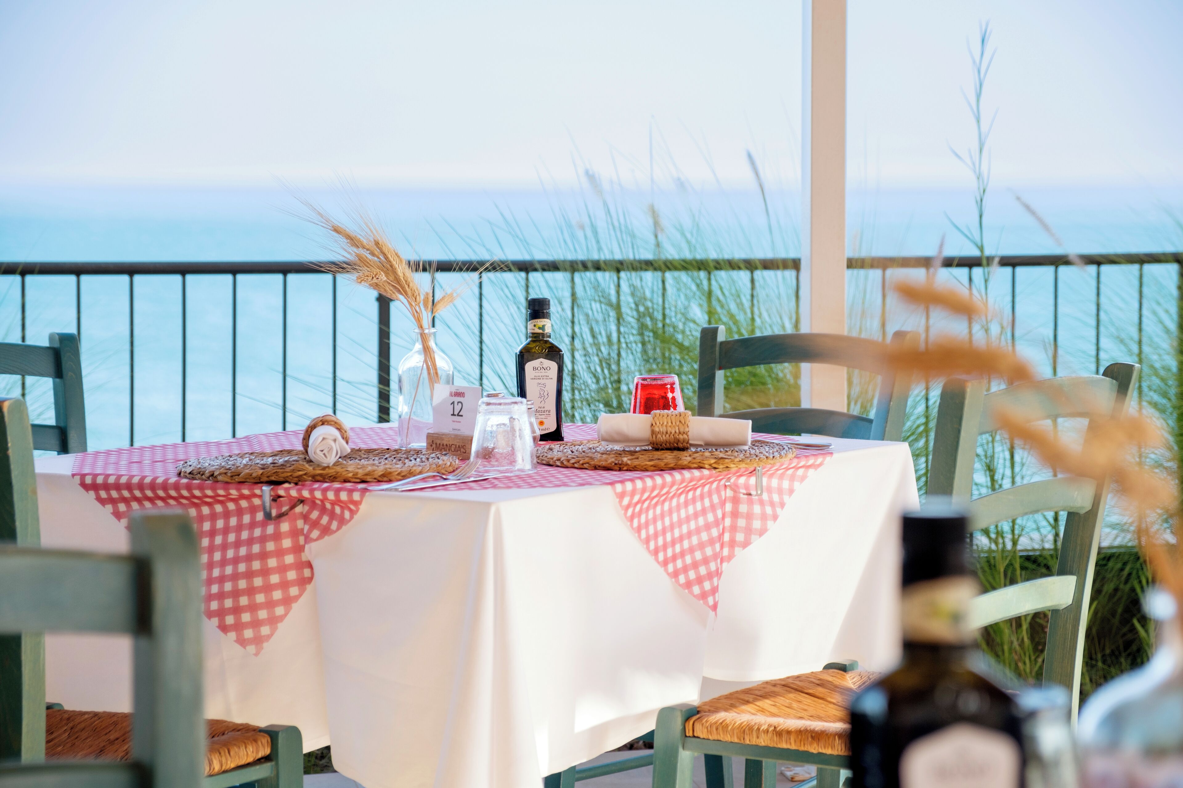 A table set for dining with a view of the sea, featuring a checkered tablecloth, wicker placemats, and a bottle of wine.