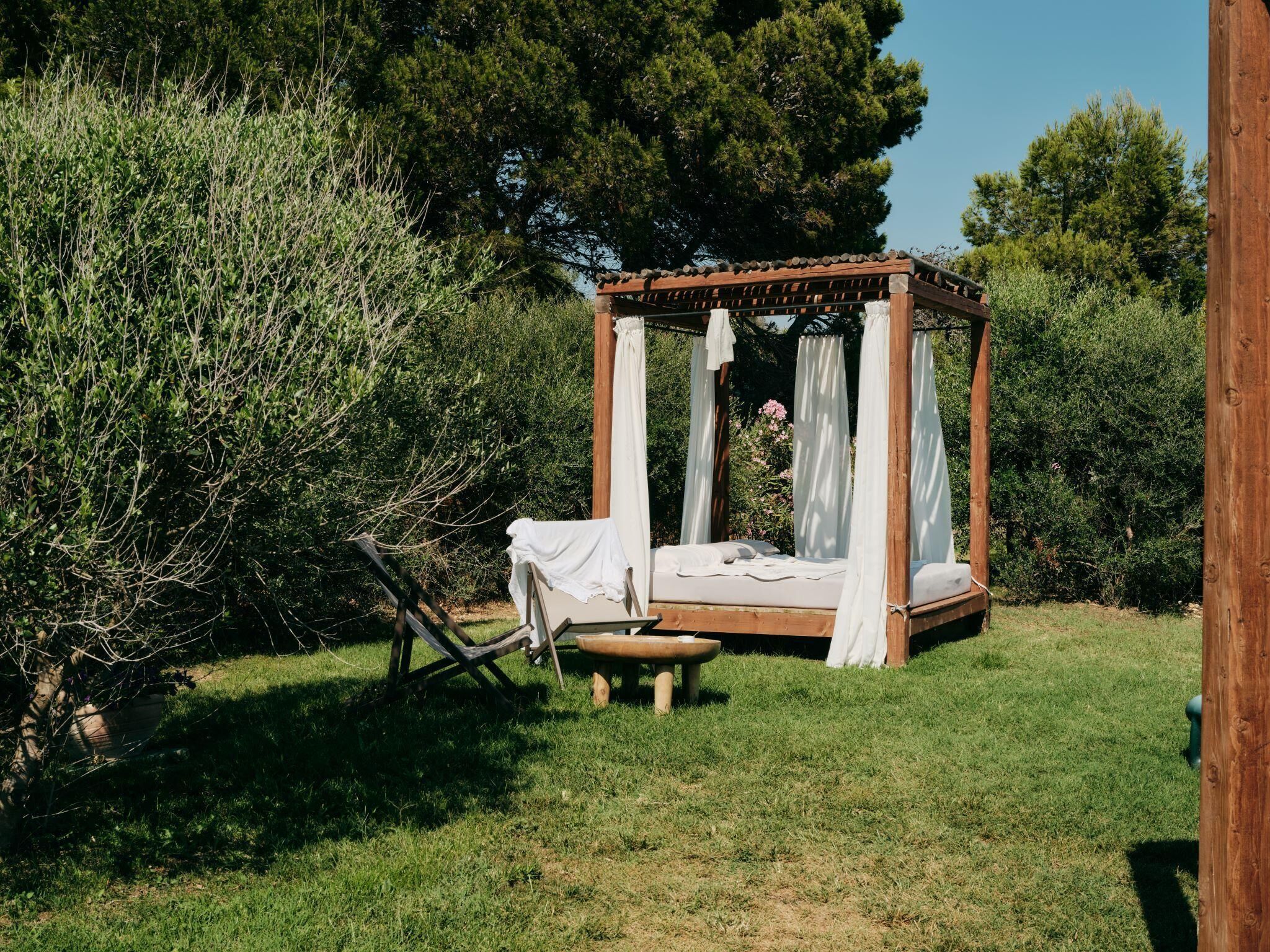Un lit à baldaquin en bois, avec des draps blancs et un banc, entouré d’une végétation luxuriante et d’arbres.