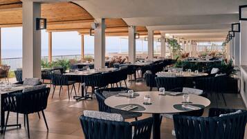Un espace de restauration en plein air avec des tables et des chaises rondes, de grandes colonnes et une vue sur la mer.