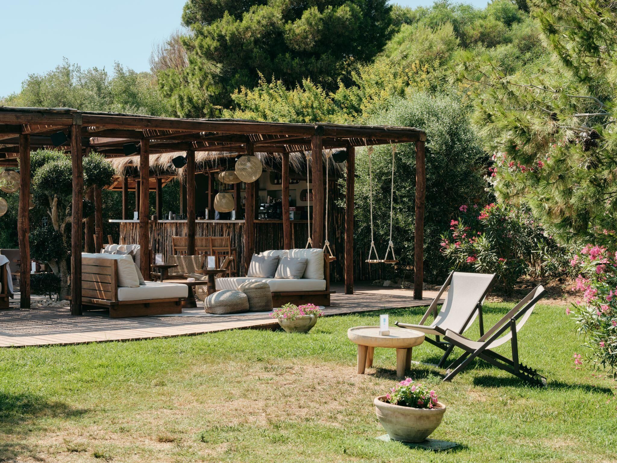 A wooden pergola with a sofa and chairs, surrounded by greenery and flowers.