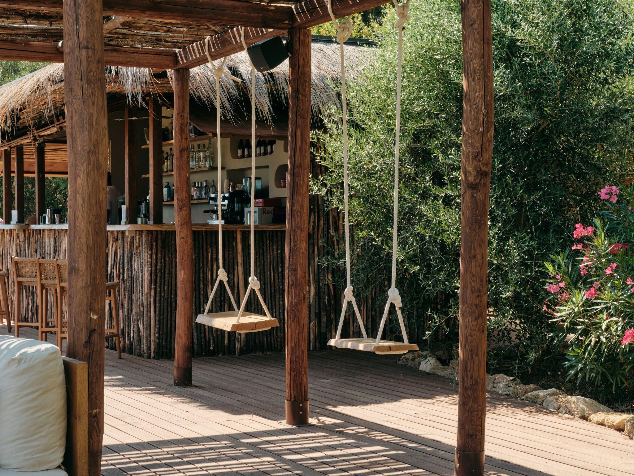 A wooden deck with hanging swings, a thatched-roof structure, and a bar area with shelves of bottles.