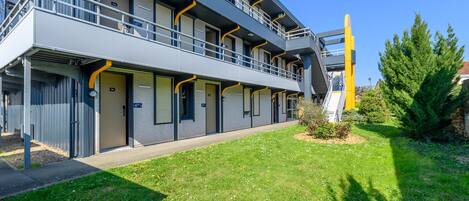 A modern building with a flat roof, multiple balconies, and a small garden area with grass and flowers.