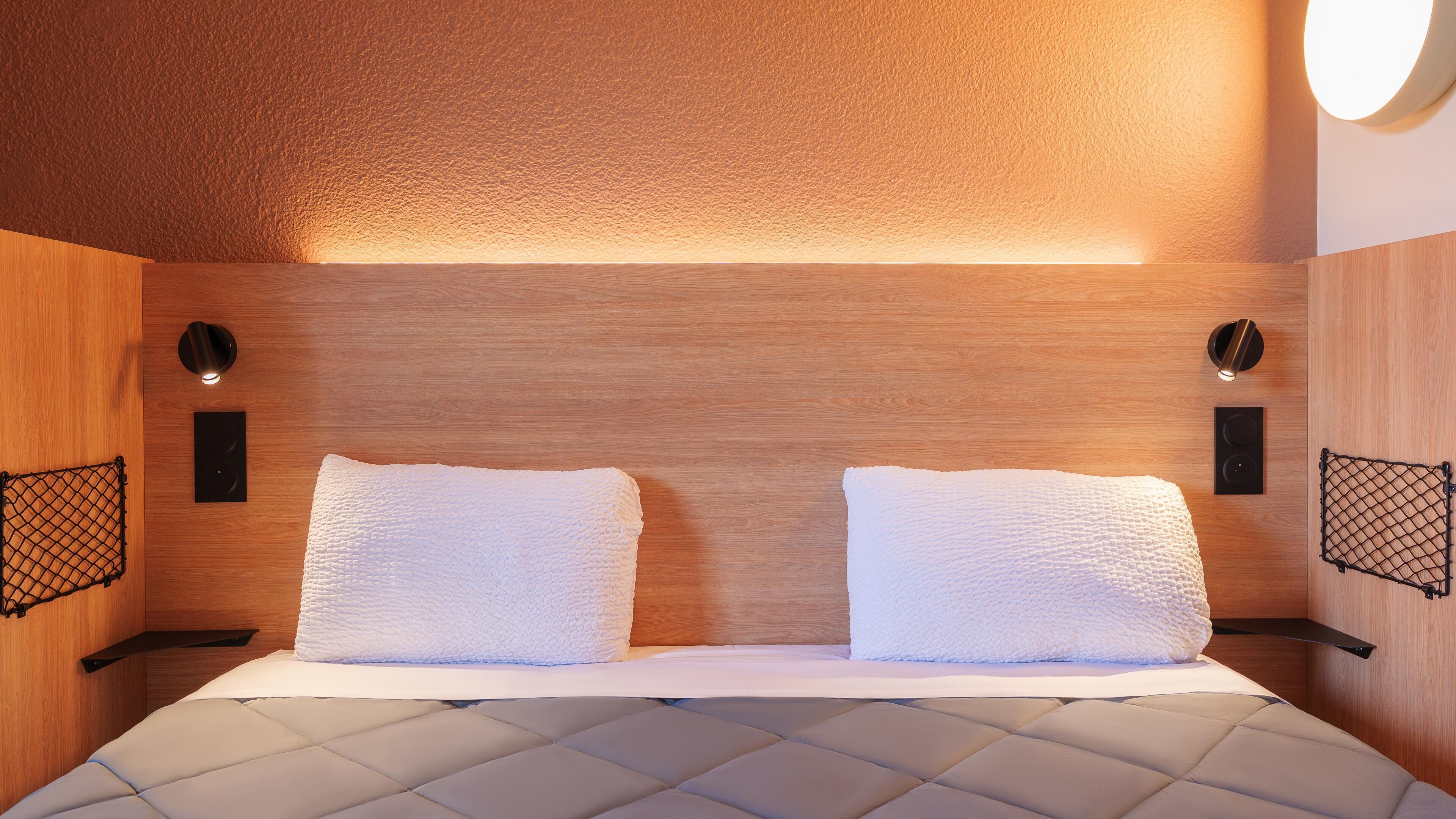 A modern hotel room with a wooden headboard, two white pillows, a quilted bedspread, and wall-mounted speakers.