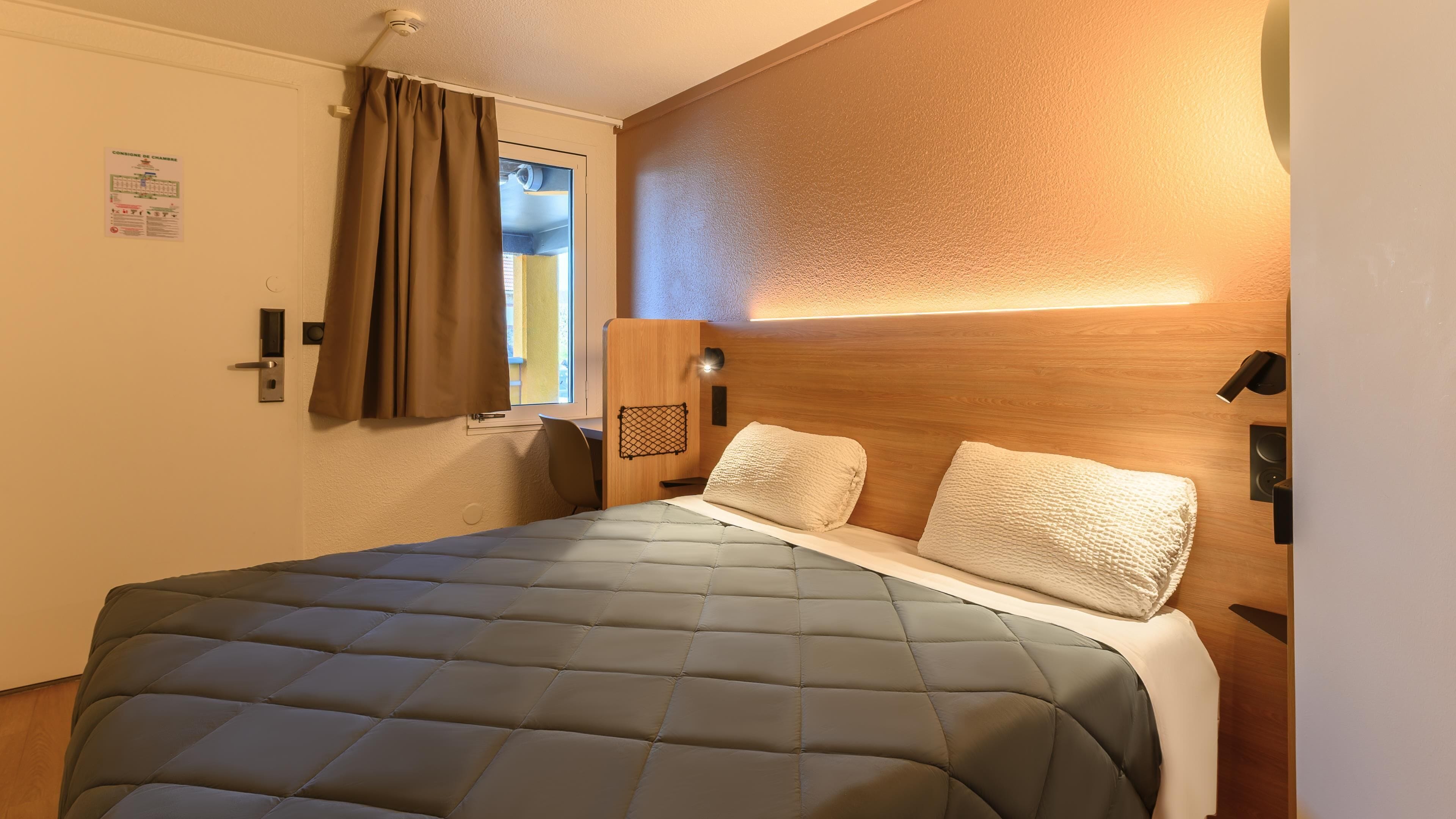 Een slaapkamer met een bed, een raam met gordijnen, een houten hoofdeinde en een wandlamp.