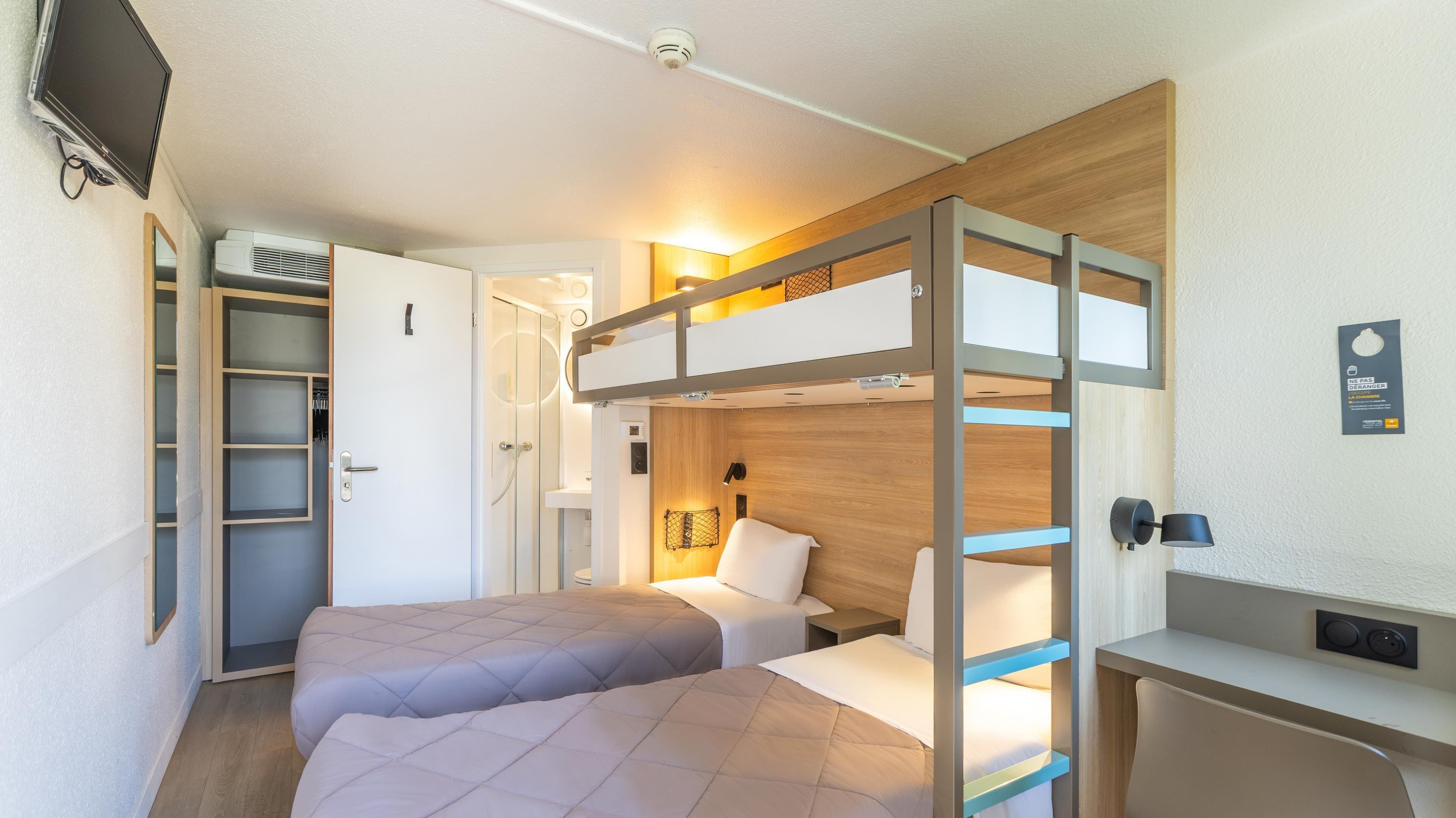 A compact hotel room with a bunk bed, a desk, a small bathroom, and a TV mounted on the wall.