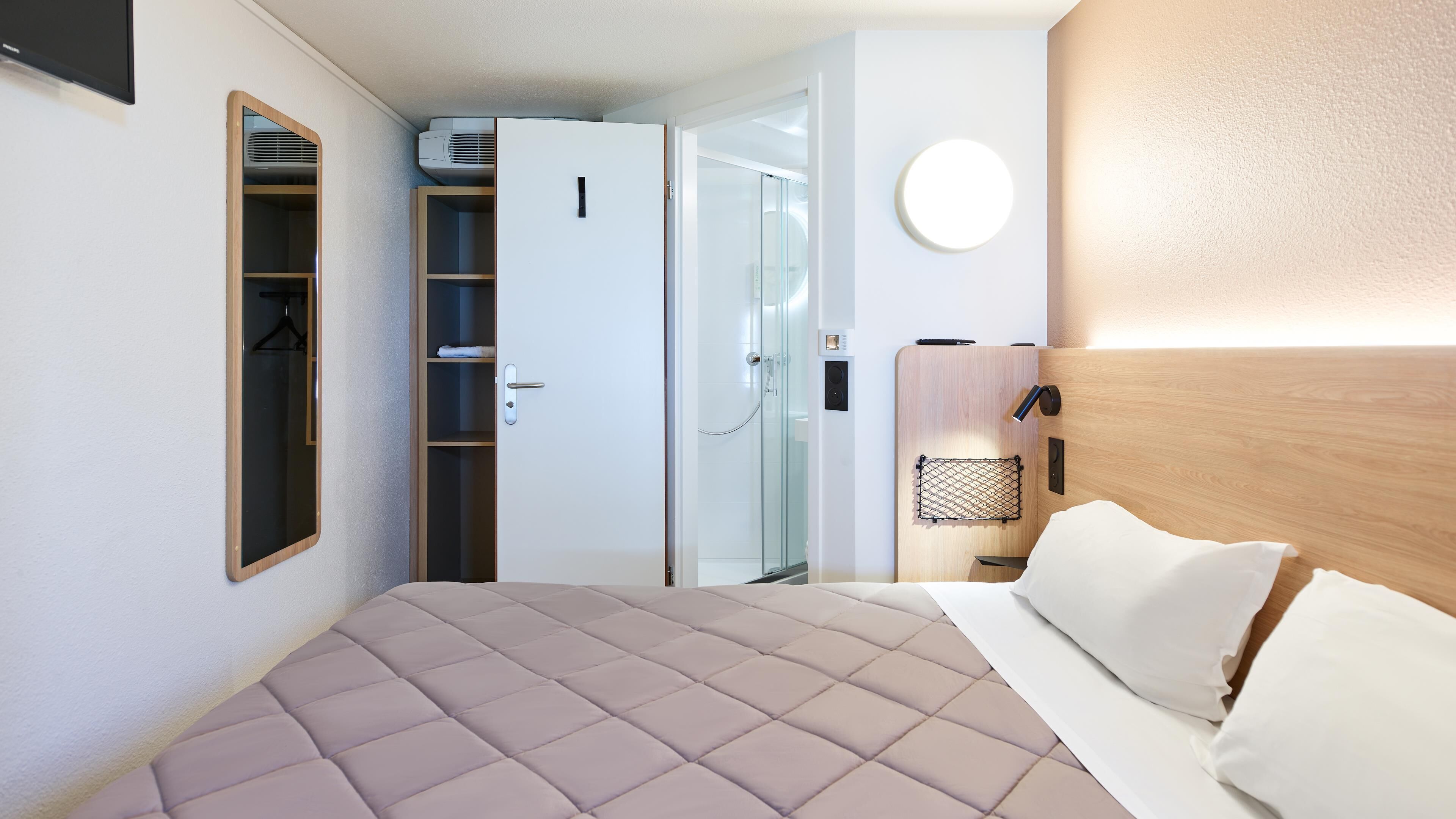 A modern hotel room with a bed, a wardrobe, a shower, and a bedside table with a lamp.