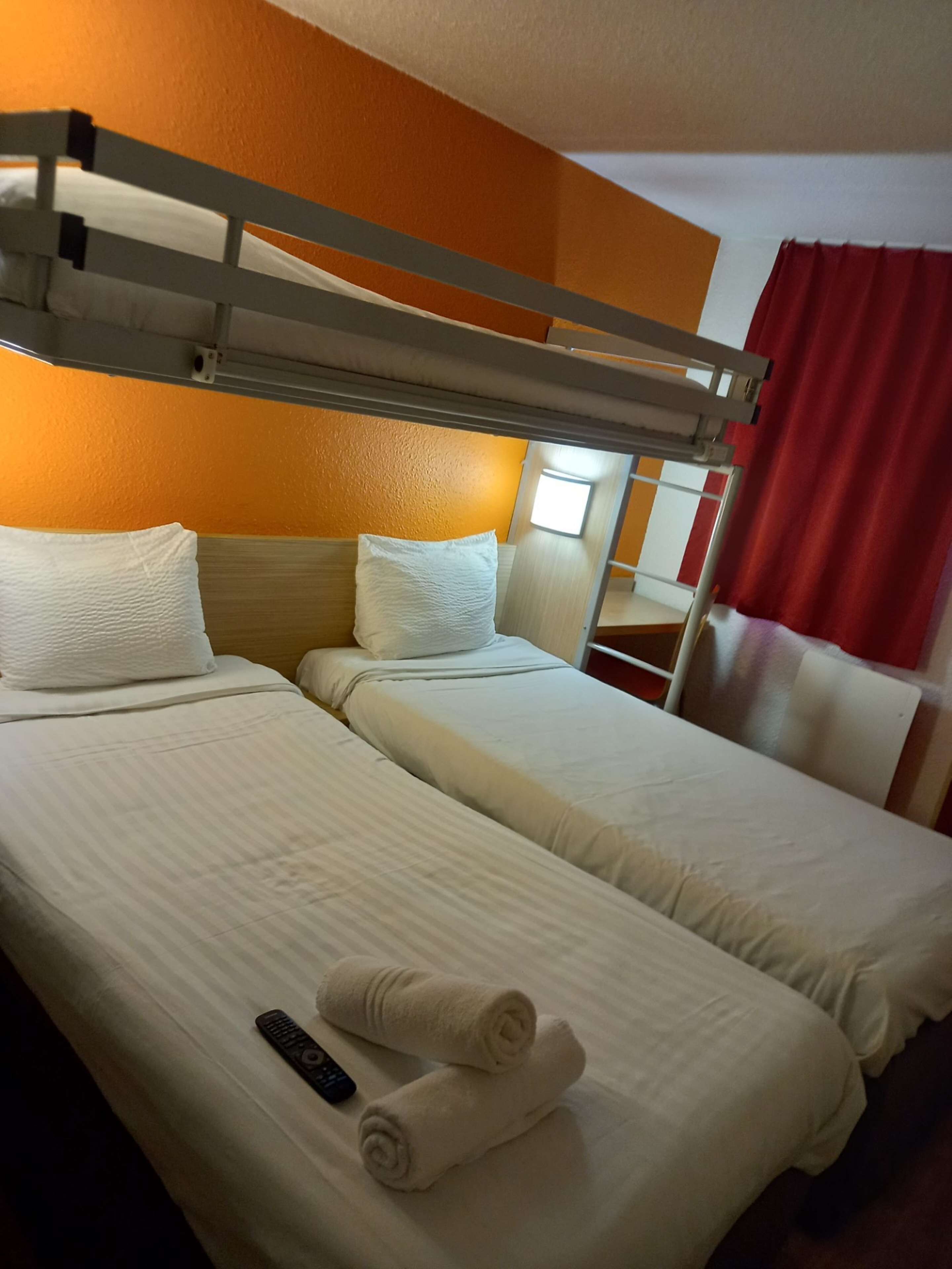 A hotel room with two beds, a bunk bed, white linens, a remote control, and a red curtain.