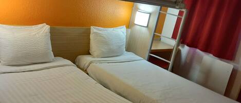 A hotel room with two beds, a bunk bed, white linens, a remote control, and a red curtain.