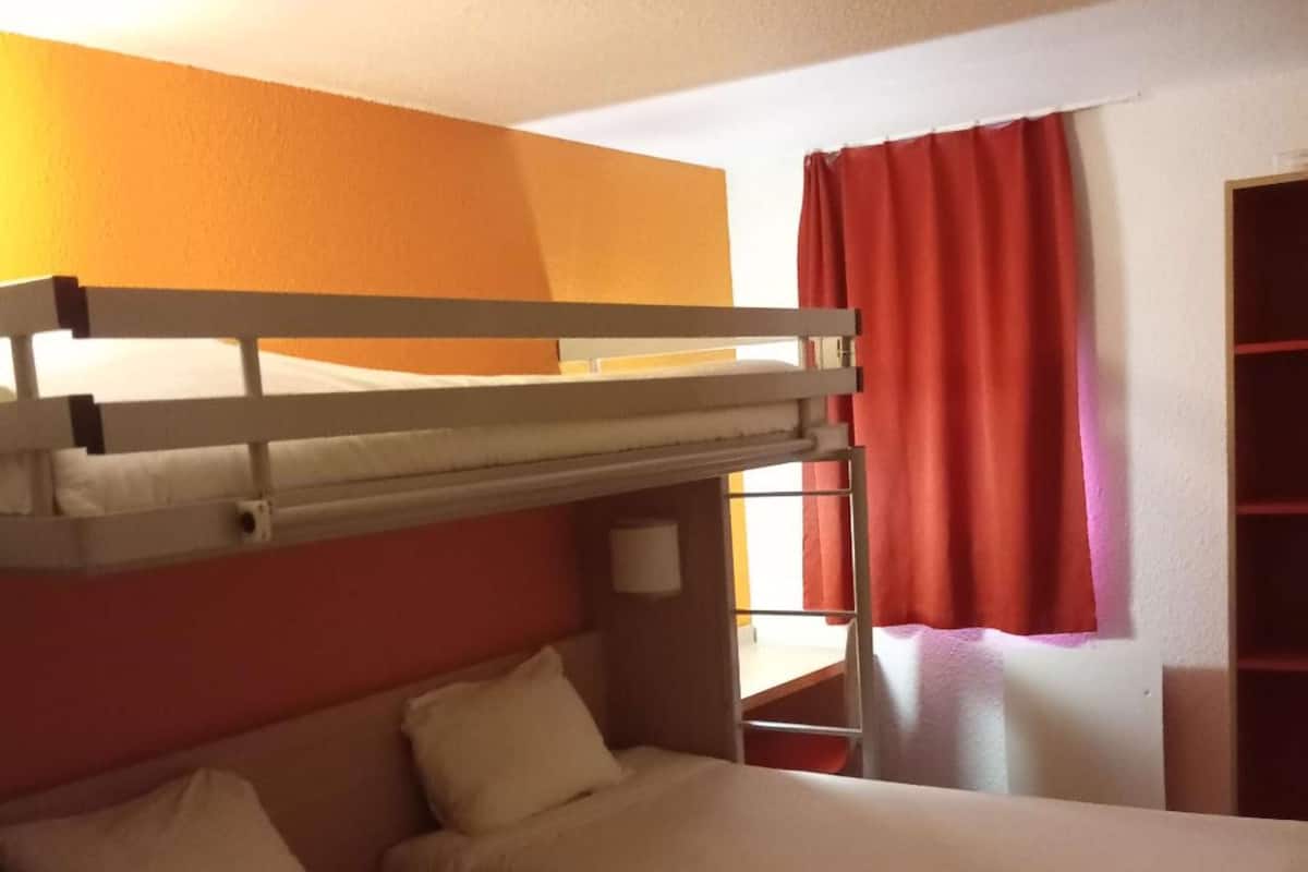 A hotel room with two beds, a bunk bed, a window with red curtains, and a small table with a lamp.