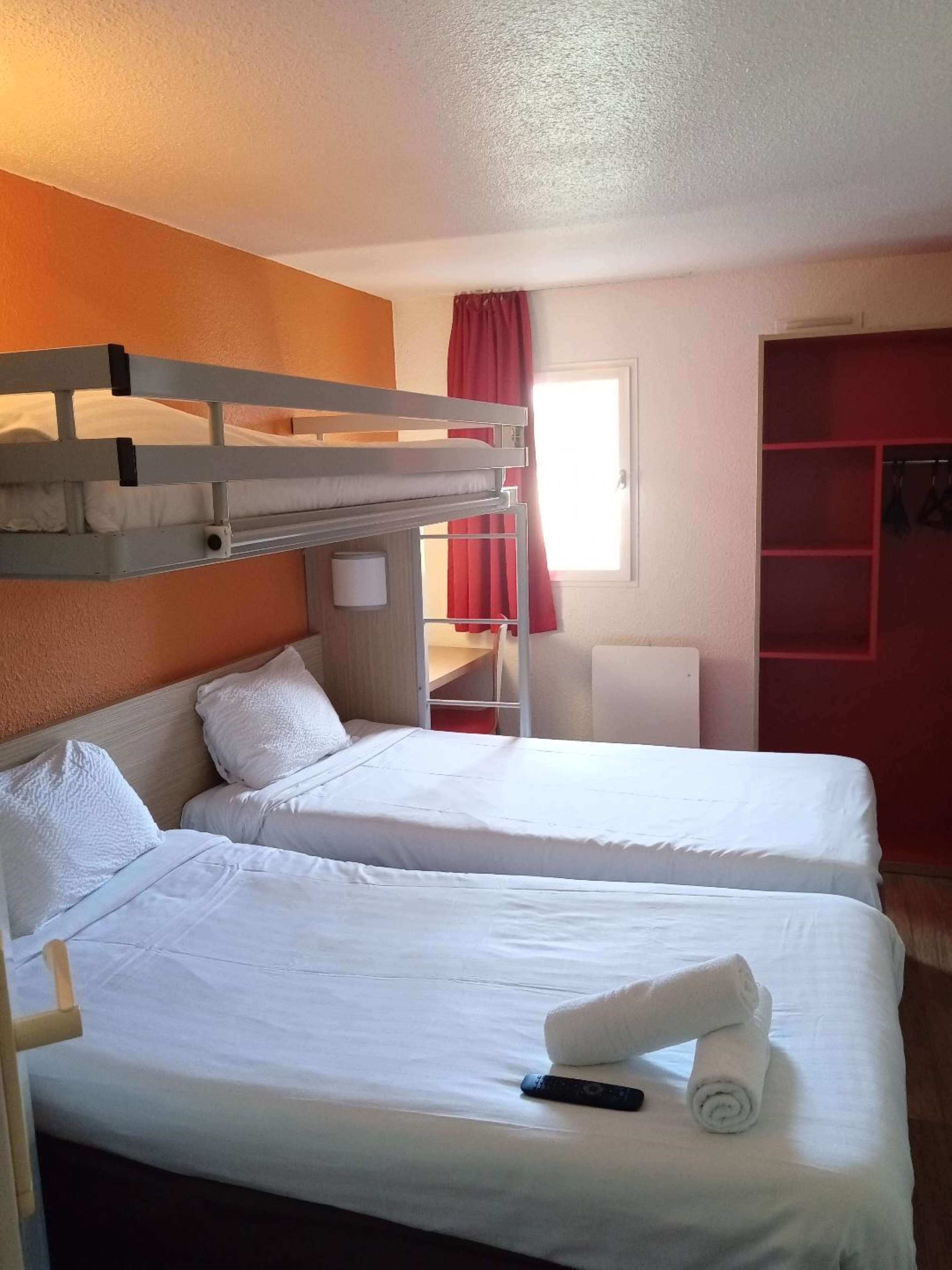 A room with two beds, a bunk bed above, a window with red curtains, and a red storage unit.