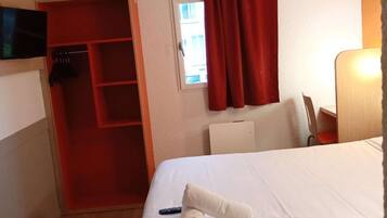 A hotel room with a bed, a TV mounted on the wall, a red wardrobe, and a window with red curtains.
