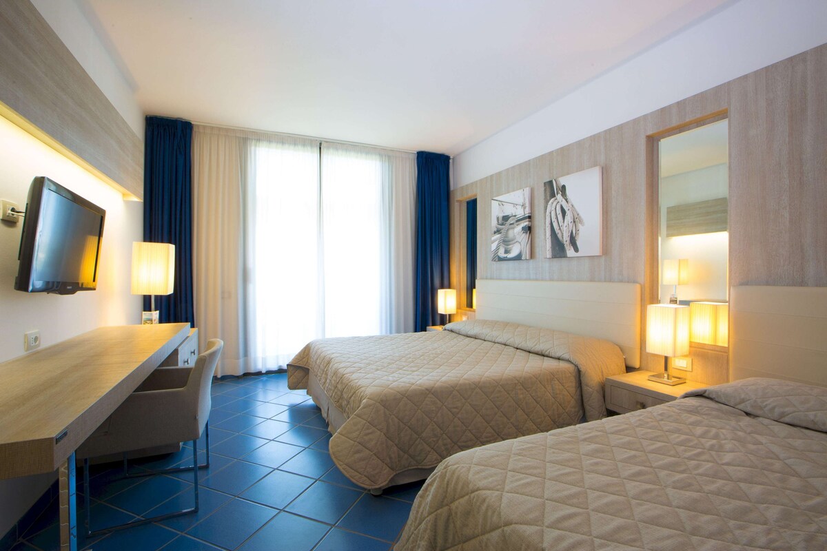A hotel room with two beds, a desk, a TV, and a large window with curtains.
