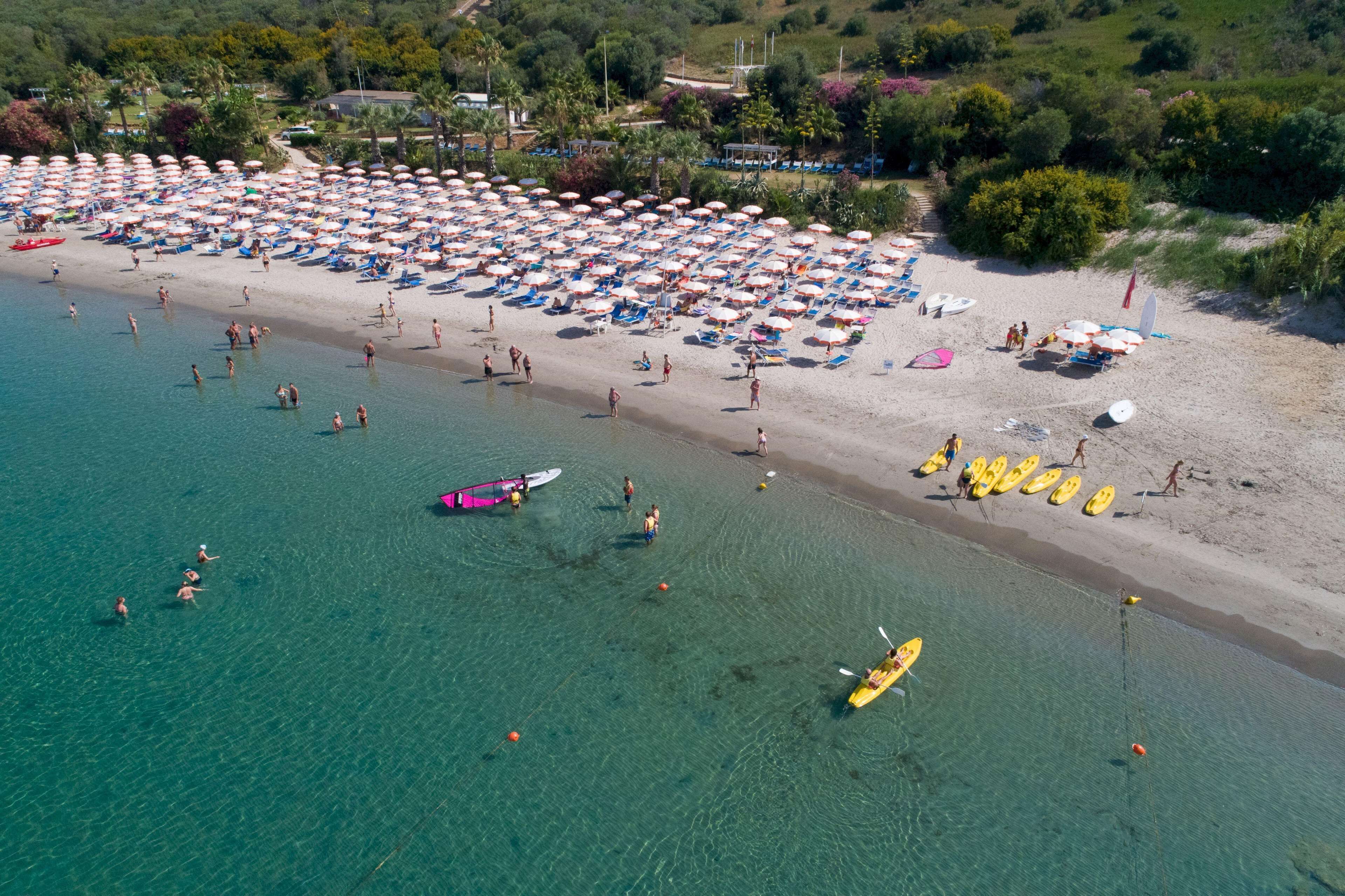 An aerial view of a beach with numerous umbrellas and beach chairs, a sandy shore, and clear blue water.