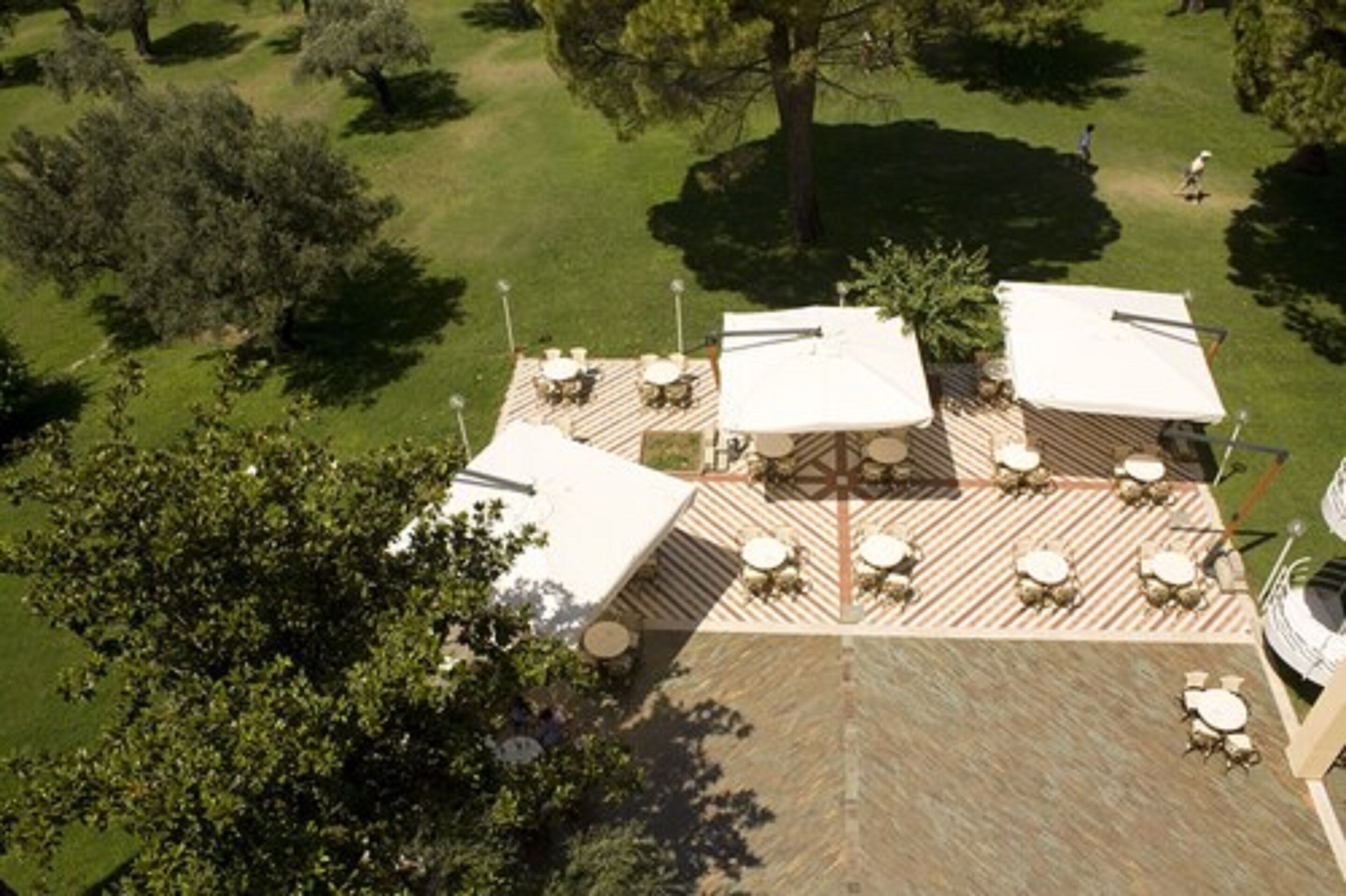 An outdoor patio area with multiple tables and umbrellas, surrounded by trees and a grassy lawn.