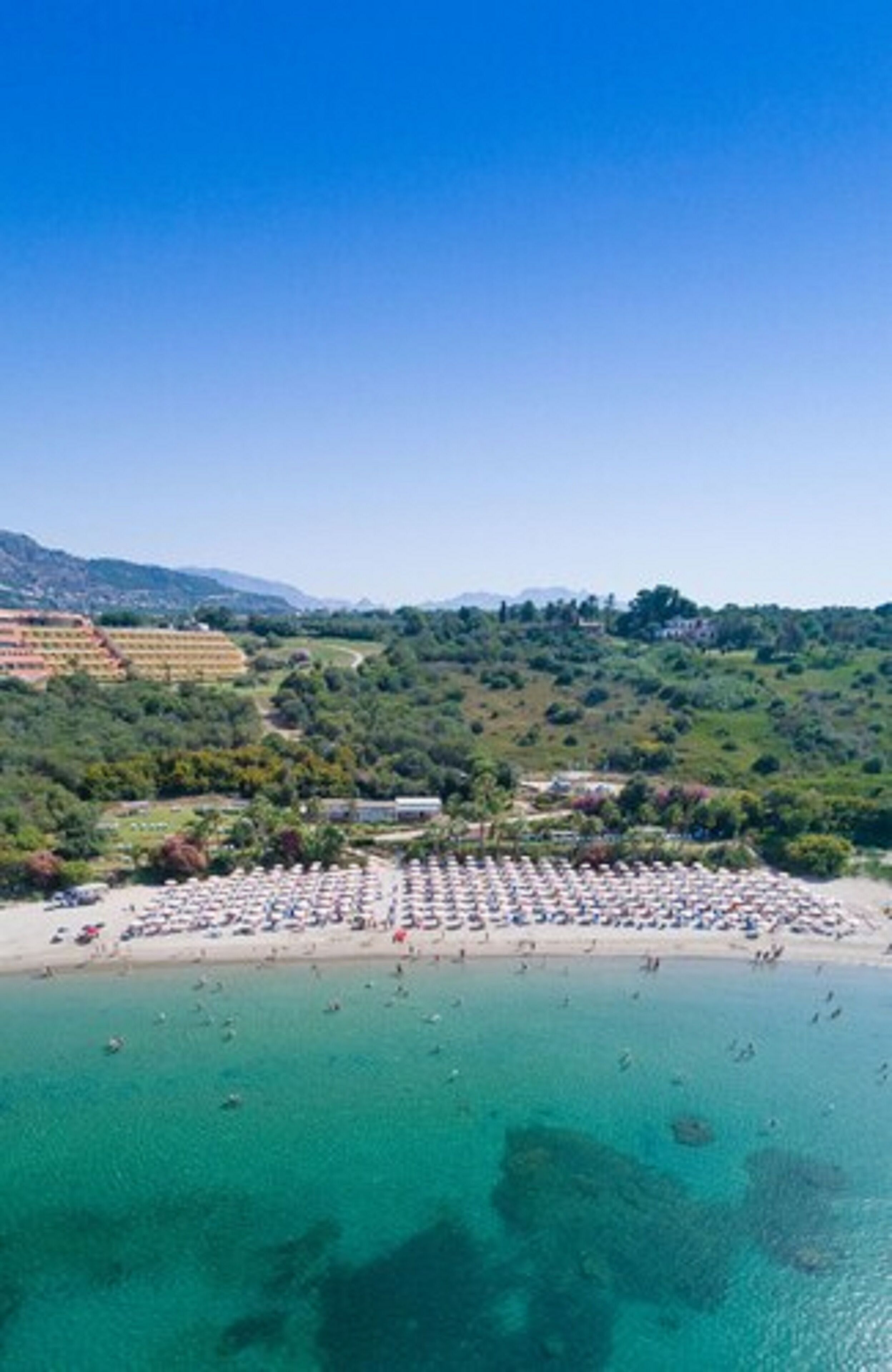 Vista aerea di una spiaggia con acque turchesi cristalline, una spiaggia sabbiosa e numerosi ombrelloni e lettini.