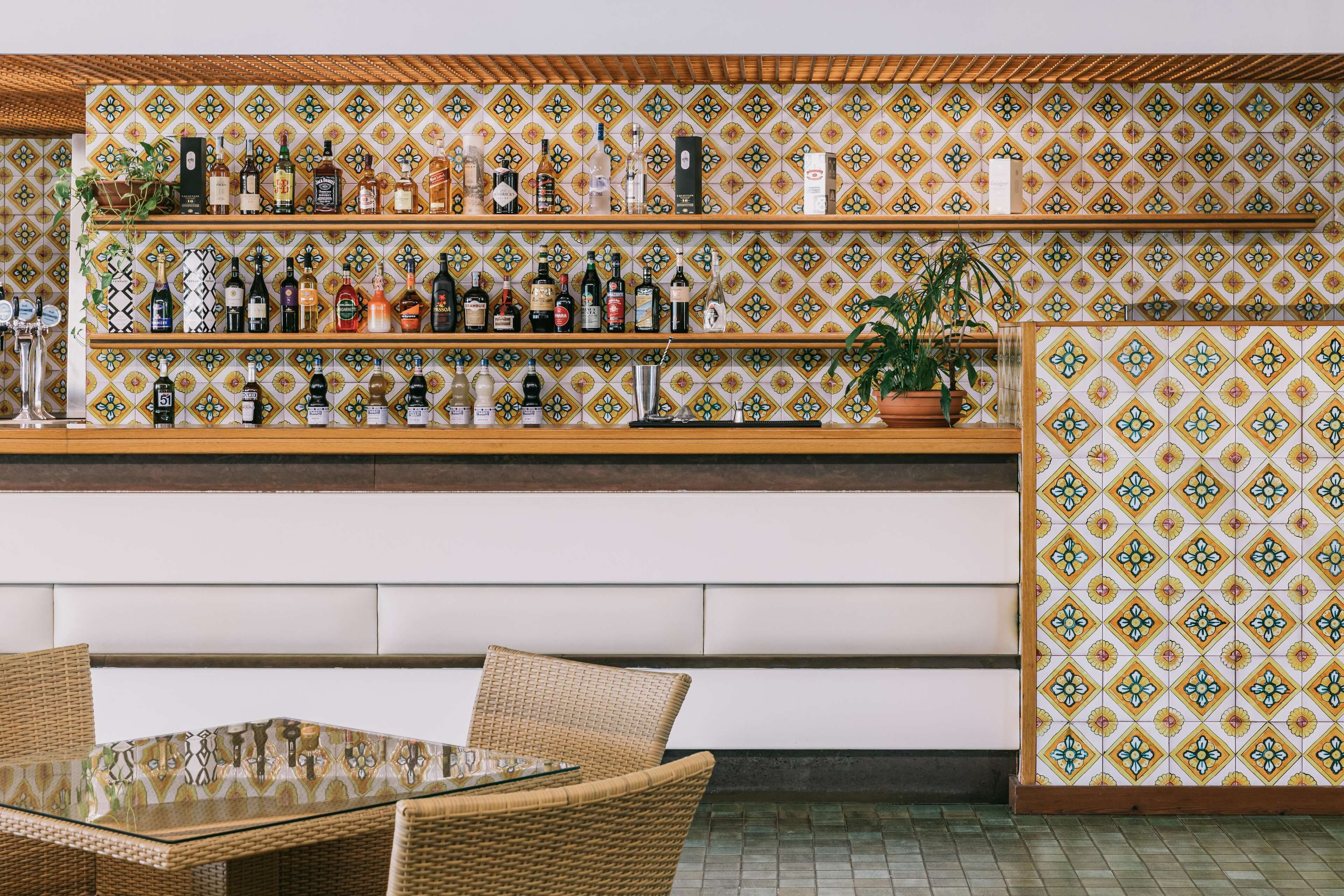 Una zona de bar con estanterías llenas de botellas de licor, mobiliario de mimbre y paredes con baldosas estampadas.