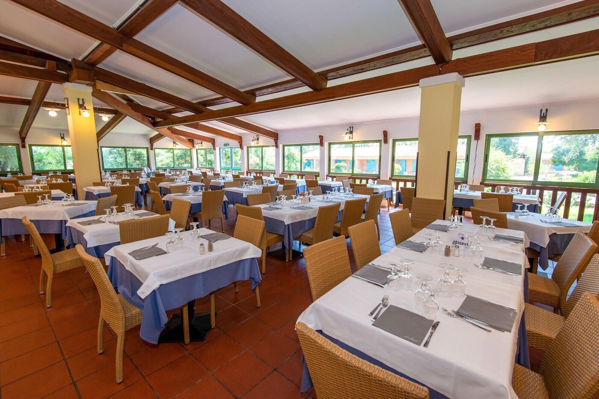 A spacious dining area with multiple tables set for guests, featuring wicker chairs and a high ceiling with wooden beams.