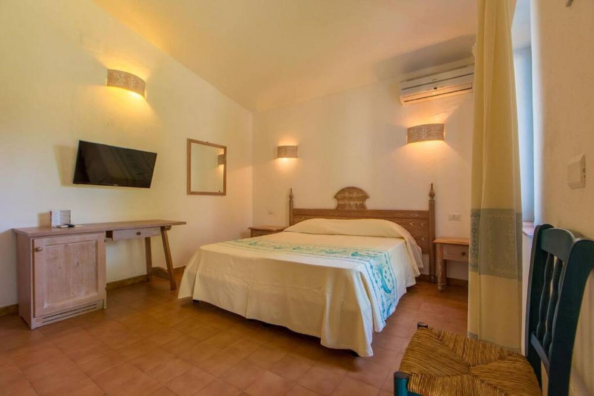 A bedroom with a bed, bedside tables, a desk, a television, and wall-mounted lights.