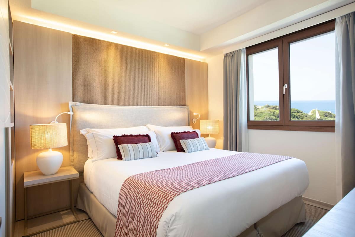 Habitación de hotel con una cama grande, dos lámparas de noche, vista al mar y una ventana con cortinas.