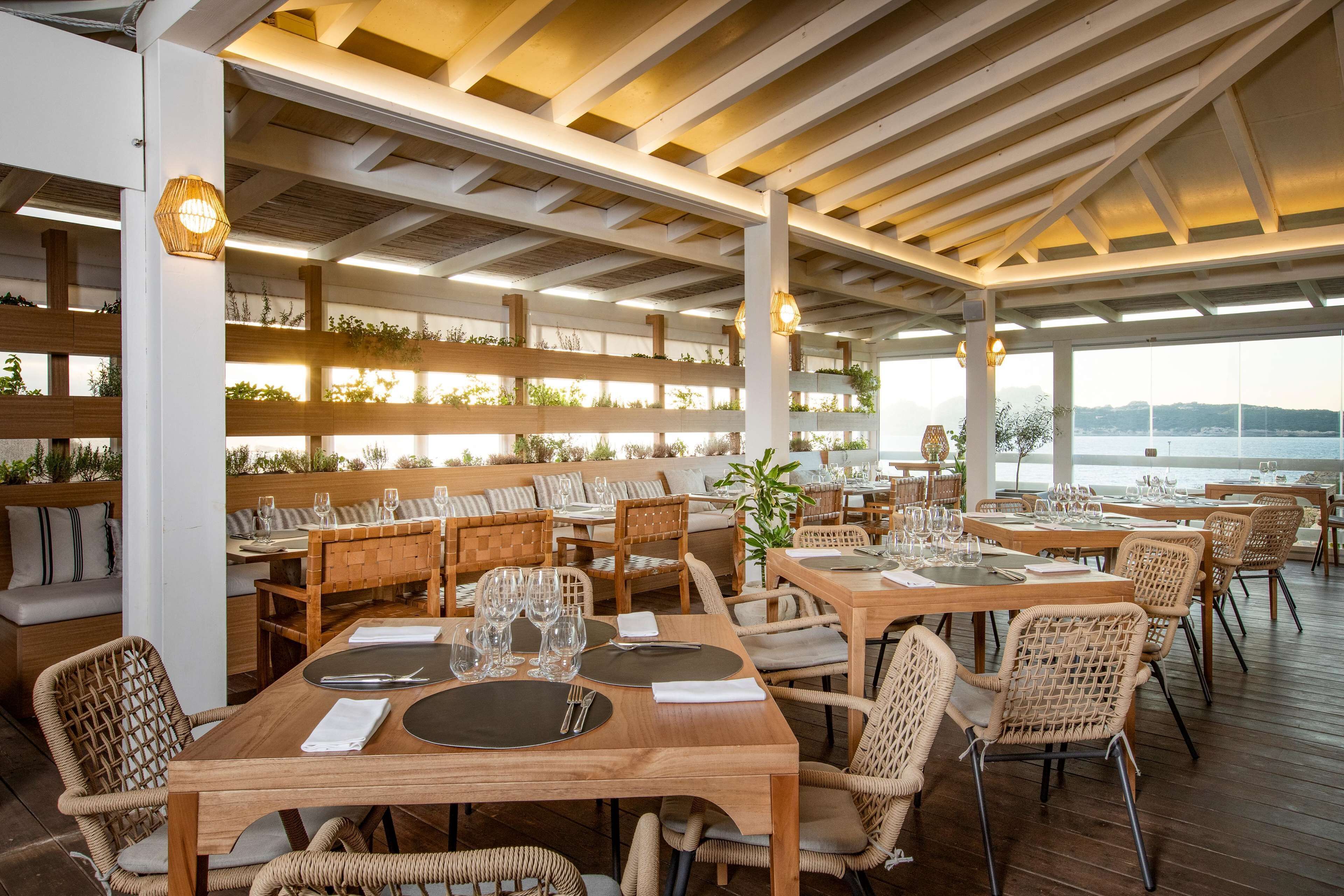 Un ristorante con tavoli di legno e sedie in vimini, soffitto alto con travi a vista e vista sul mare.