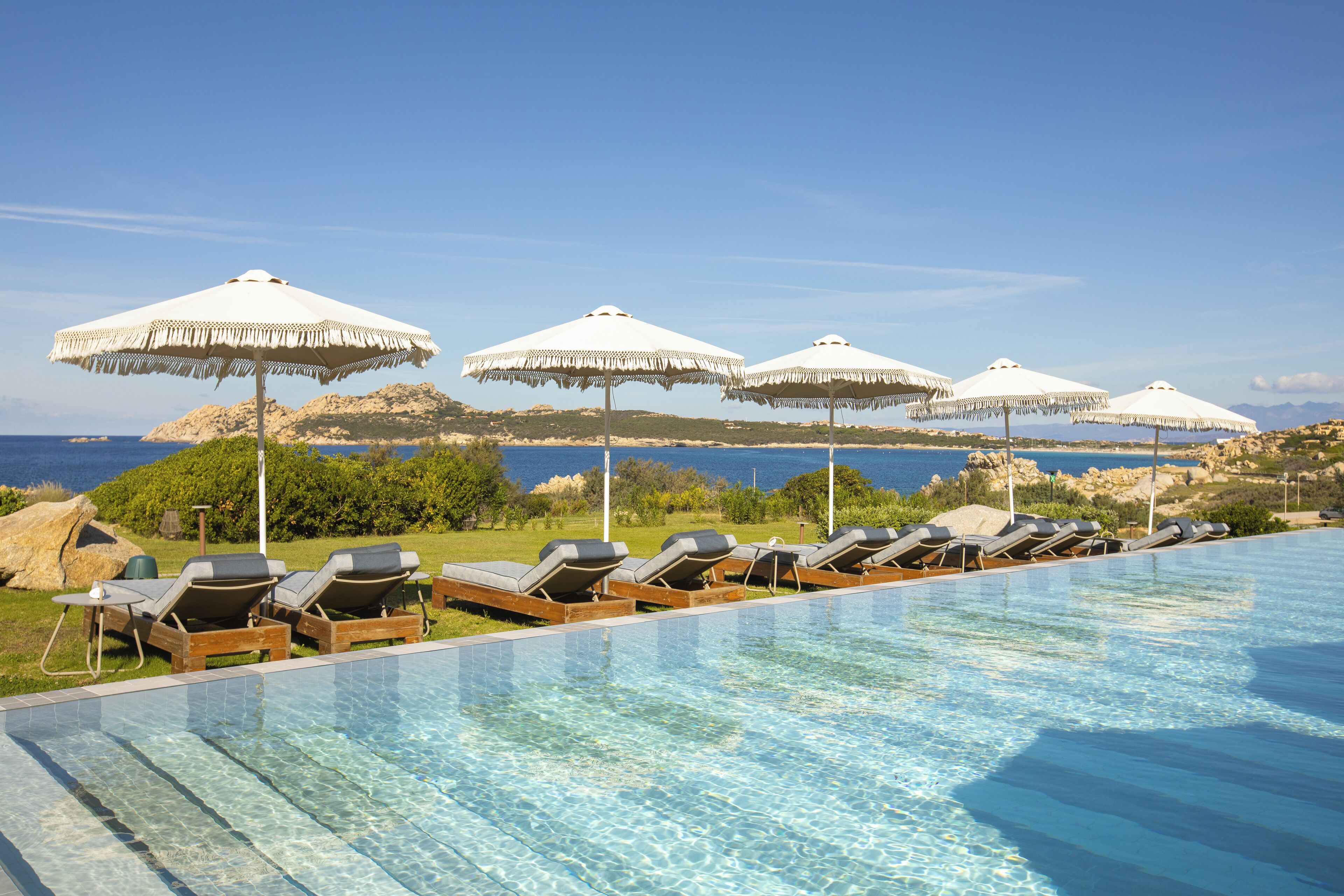 Ein Poolbereich mit Liegestühlen und Sonnenschirmen, mit Blick auf ein Gewässer und eine felsige Landschaft.