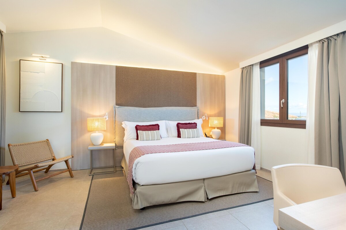 A hotel room with a large bed, two bedside lamps, a chair, and a view of the beach through the window.