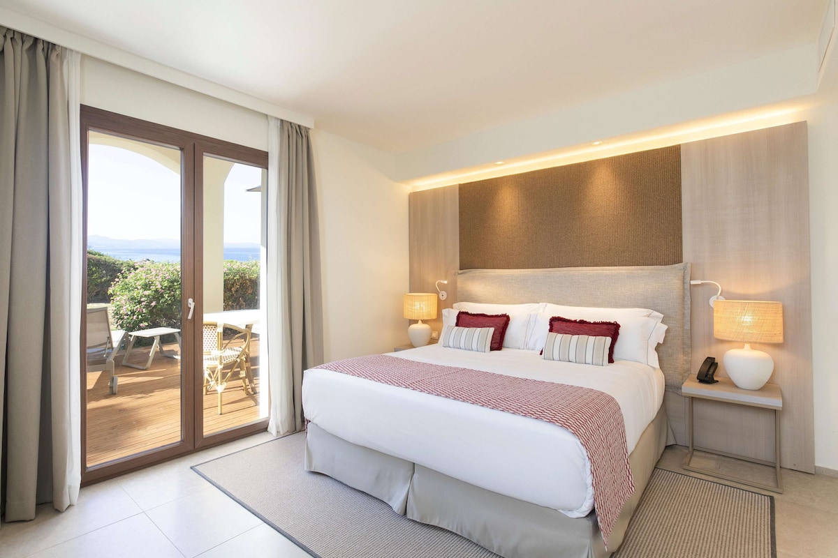 Ein Hotelzimmer mit einem großen Bett, zwei Nachttischlampen, Blick ins Freie und einem kleinen Tisch mit Stühlen.