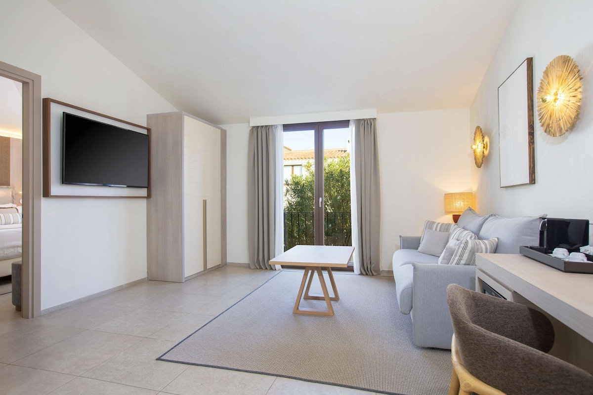 Ein modernes Hotelzimmer mit Fernseher, einer Sitzecke, einem Esstisch und einem großen Fenster mit Vorhängen.