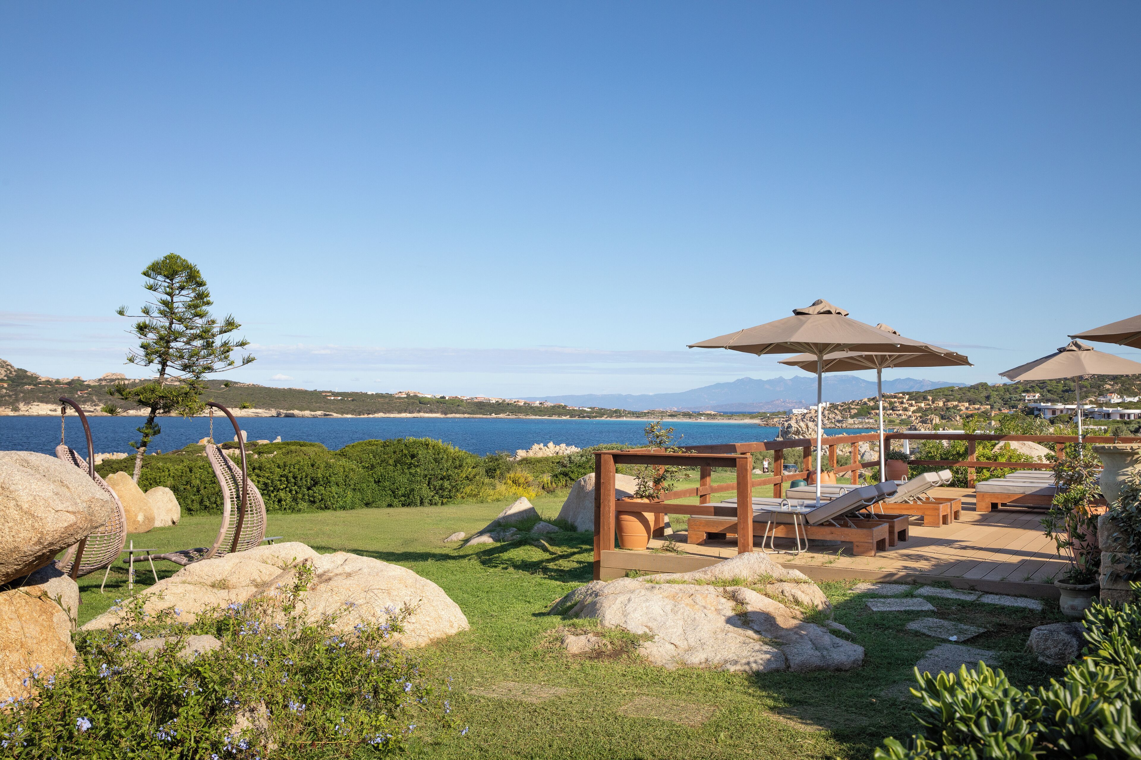 Een terras buiten aan de kust met lounge fauteuils en parasols, met uitzicht op water en heuvels.