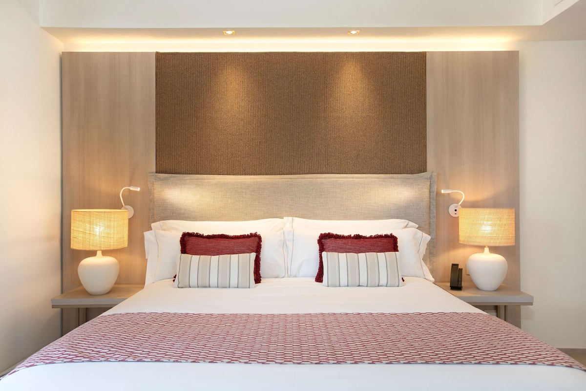 Una cama bien hecha con ropa blanca y detalles rojos, flanqueada por dos mesitas de noche con lámparas.