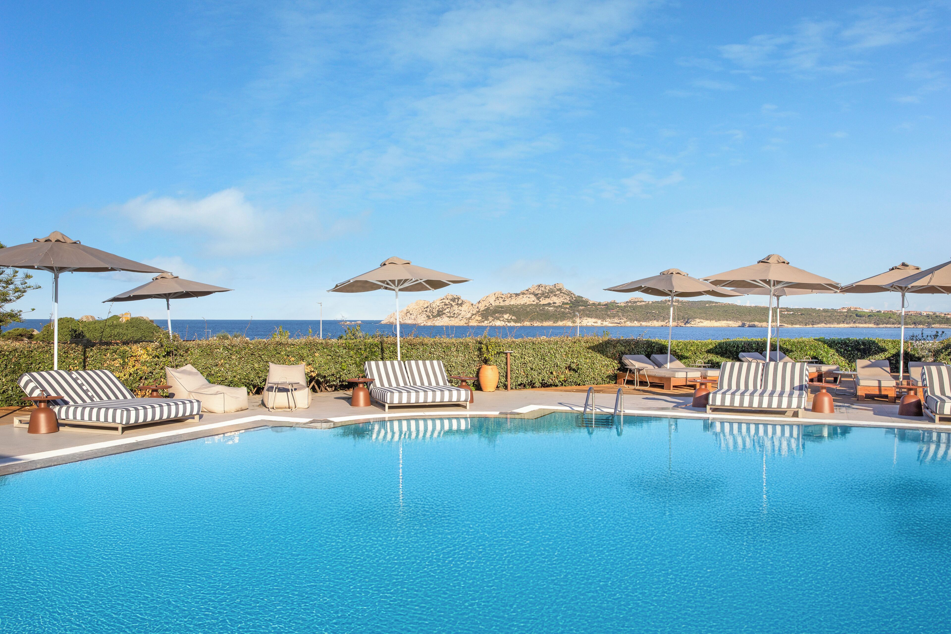 A pool with lounge chairs and umbrellas, a clear sky, and a view of the sea.
