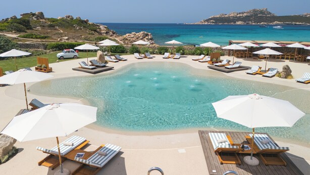 Ein Poolbereich mit Liegestühlen und Sonnenschirmen, ein klares Schwimmbecken und Blick auf das Meer und die felsige Landschaft.