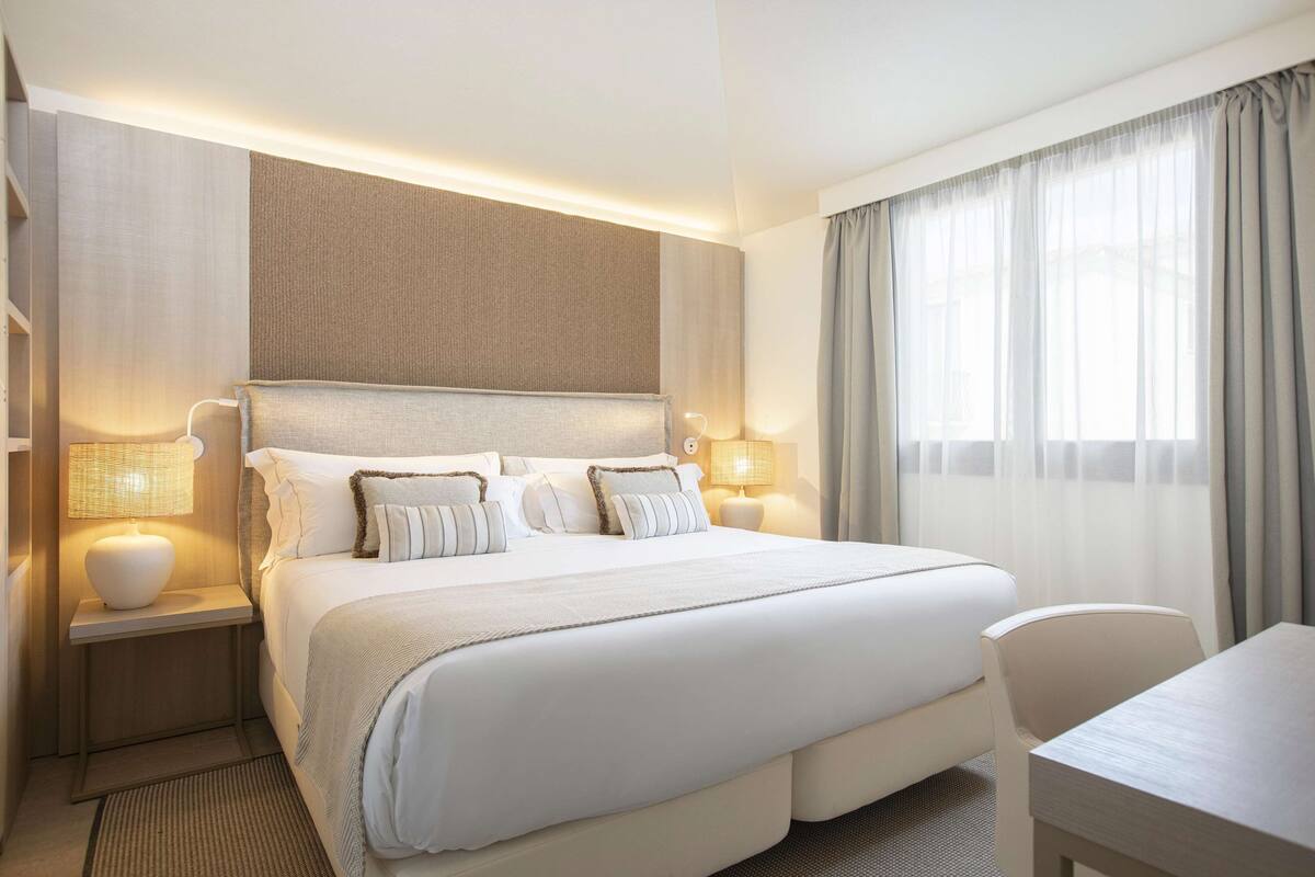 Habitación de hotel con una cama grande, lámparas de noche, un escritorio y vistas al exterior a través de cortinas translúcidas.