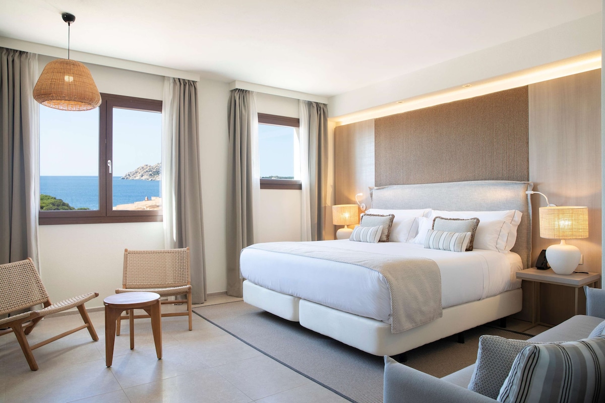 Ein Schlafzimmer mit einem großen Bett, zwei Stühlen, einem kleinen Tisch und Meerblick.