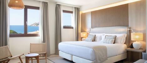 A bedroom with a large bed, two chairs, a small table, and a view of the sea.
