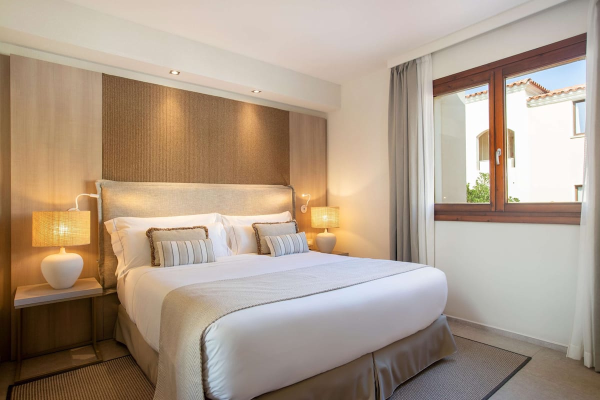 Habitación de hotel con una cama grande, dos mesitas de noche con lámparas, una ventana con cortinas y vistas a un edificio.