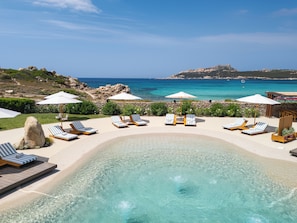 Zona de piscina con sillones de descanso y sombrillas, con vistas a una playa y al mar.