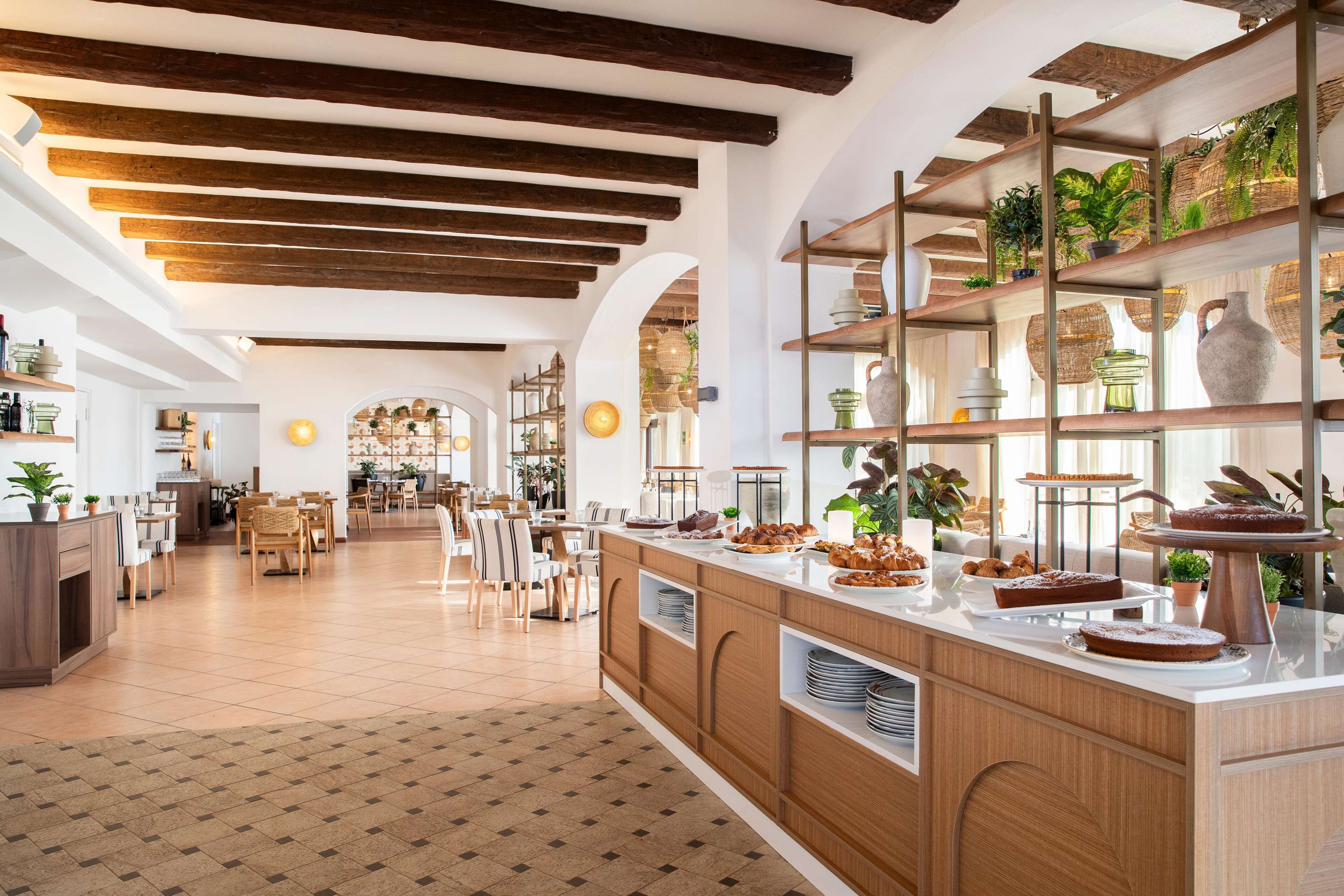 A spacious kitchen with wooden beams, a central island with a variety of food items, and shelves with plants and decorative items.