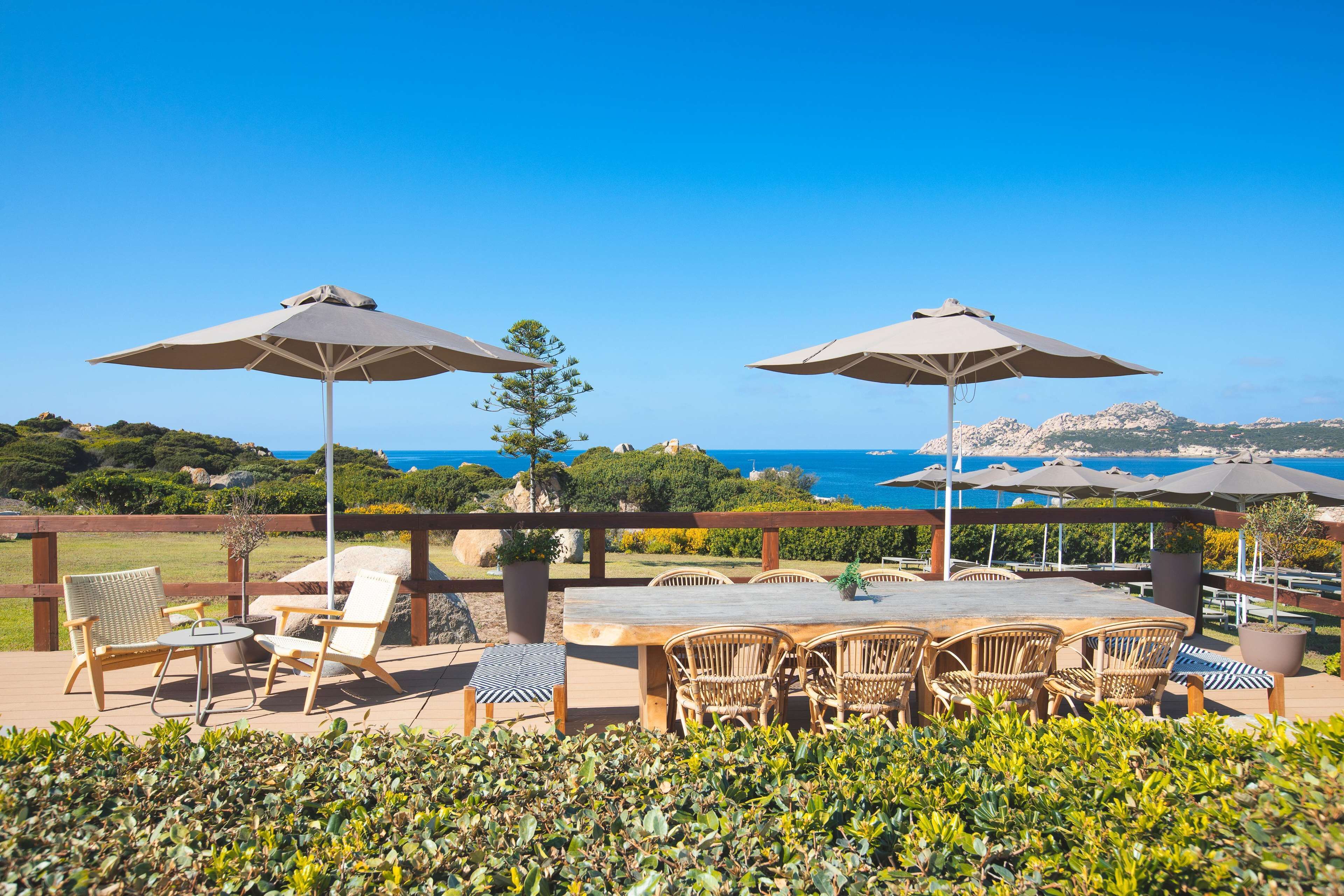 Une terrasse extérieure avec une table et des chaises en bois, deux grands parasols et une vue sur la mer.
