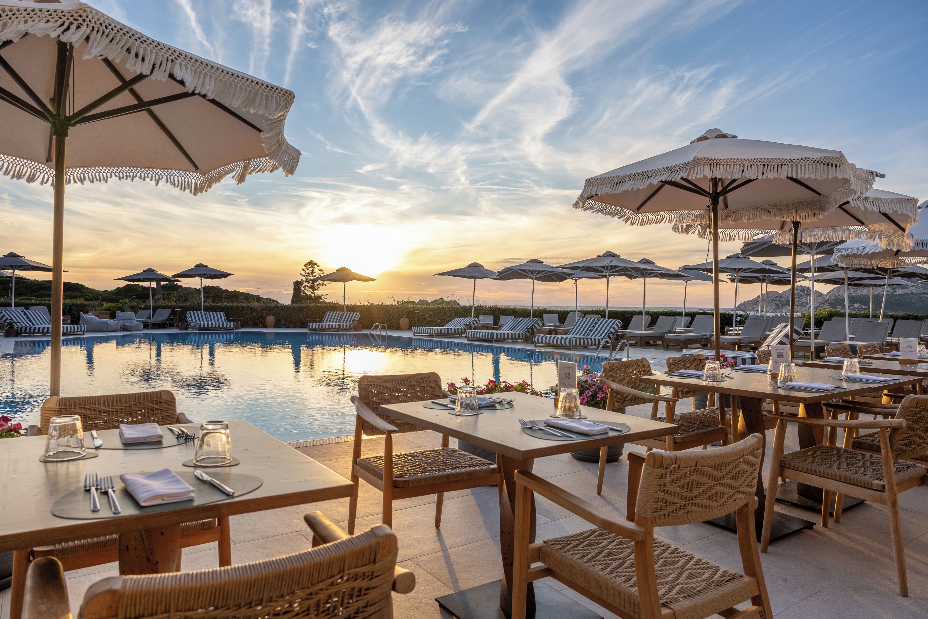 Un área de comedor junto a la piscina con mesas, sillas y sombrillas, con vistas a una piscina y tumbonas.