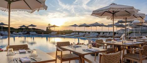 A poolside dining area with tables, chairs, and umbrellas, overlooking a swimming pool and lounge chairs.