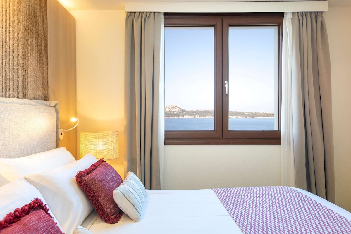 Ein Hotelzimmer mit Bett, Nachttisch, Lampe und Fenster mit Meerblick.
