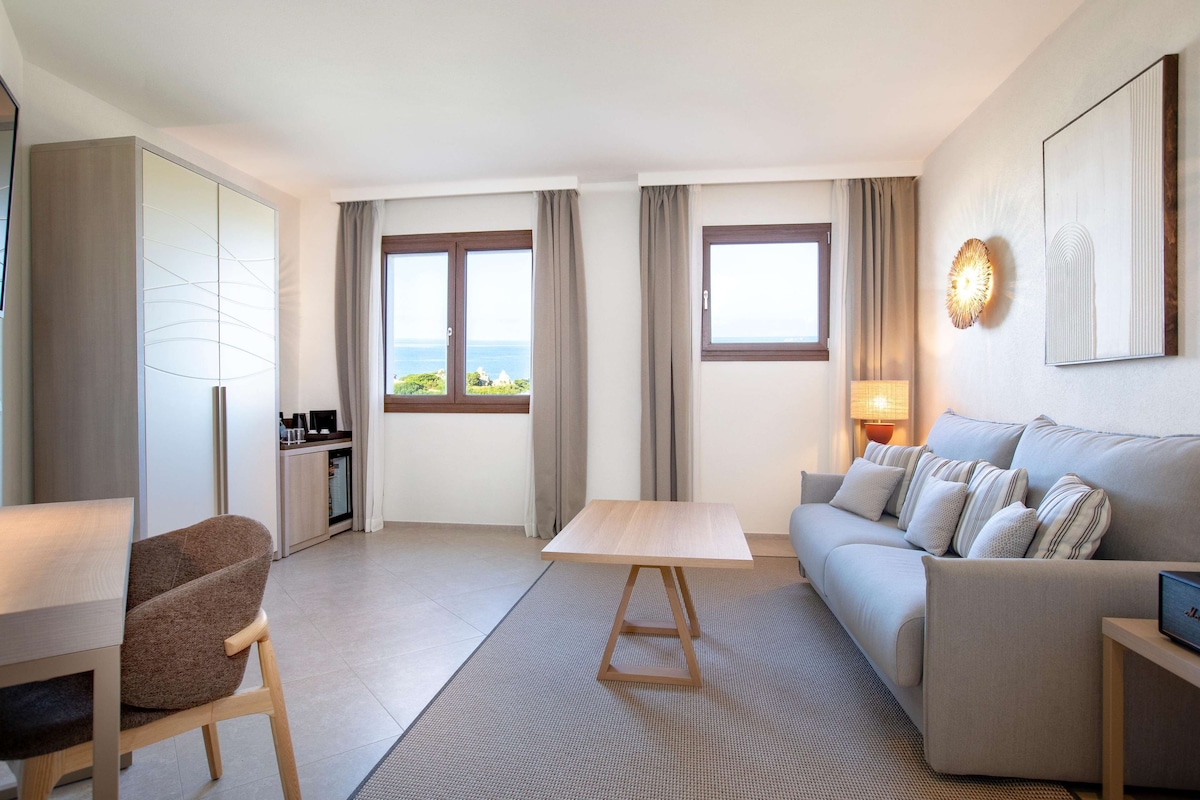 Ein modernes Hotelzimmer mit einer Couch, einem Esstisch, einem Stuhl, einem Kleiderschrank und Meerblick.