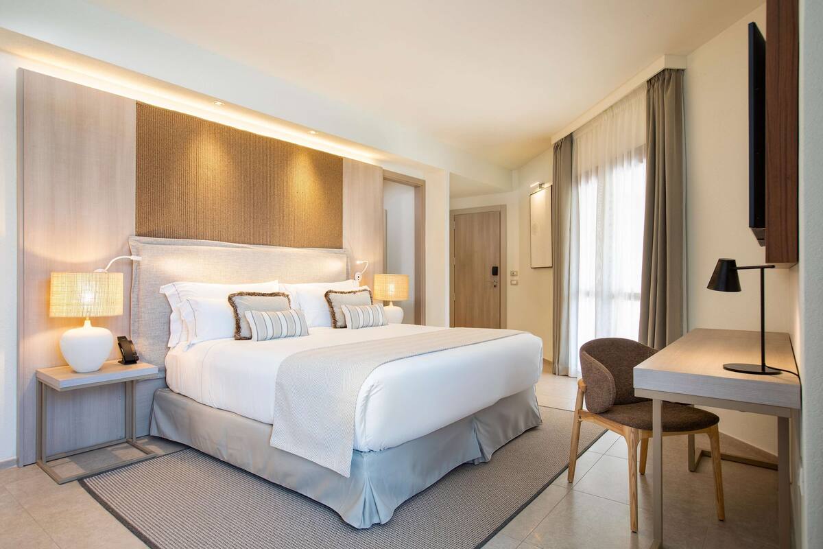 Una habitación de hotel moderna con una cama grande, mesitas de noche con lámparas, un escritorio con una silla y un televisor montado en la pared.