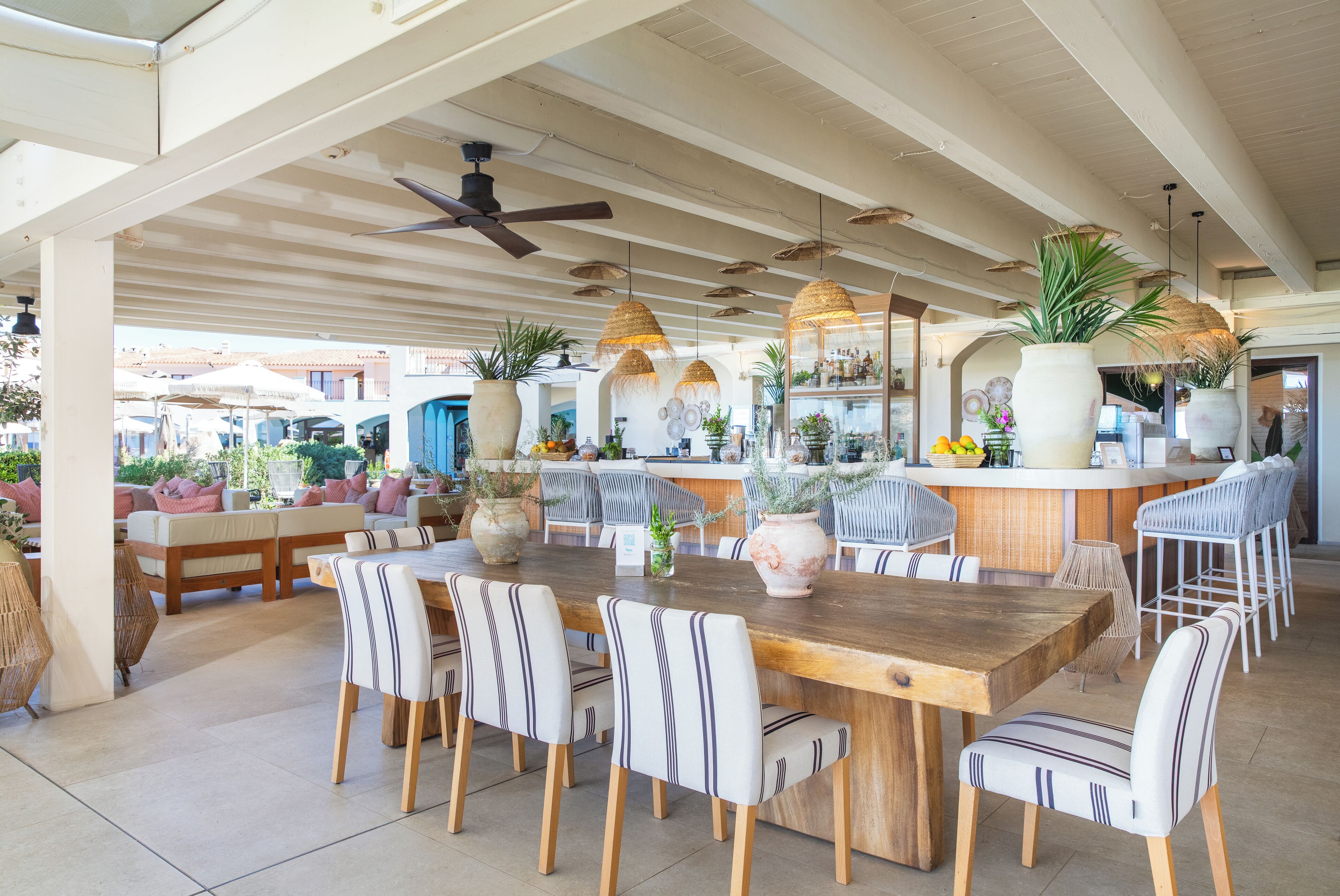 An outdoor dining area with a wooden table and striped chairs, surrounded by potted plants and a bar area with stools.