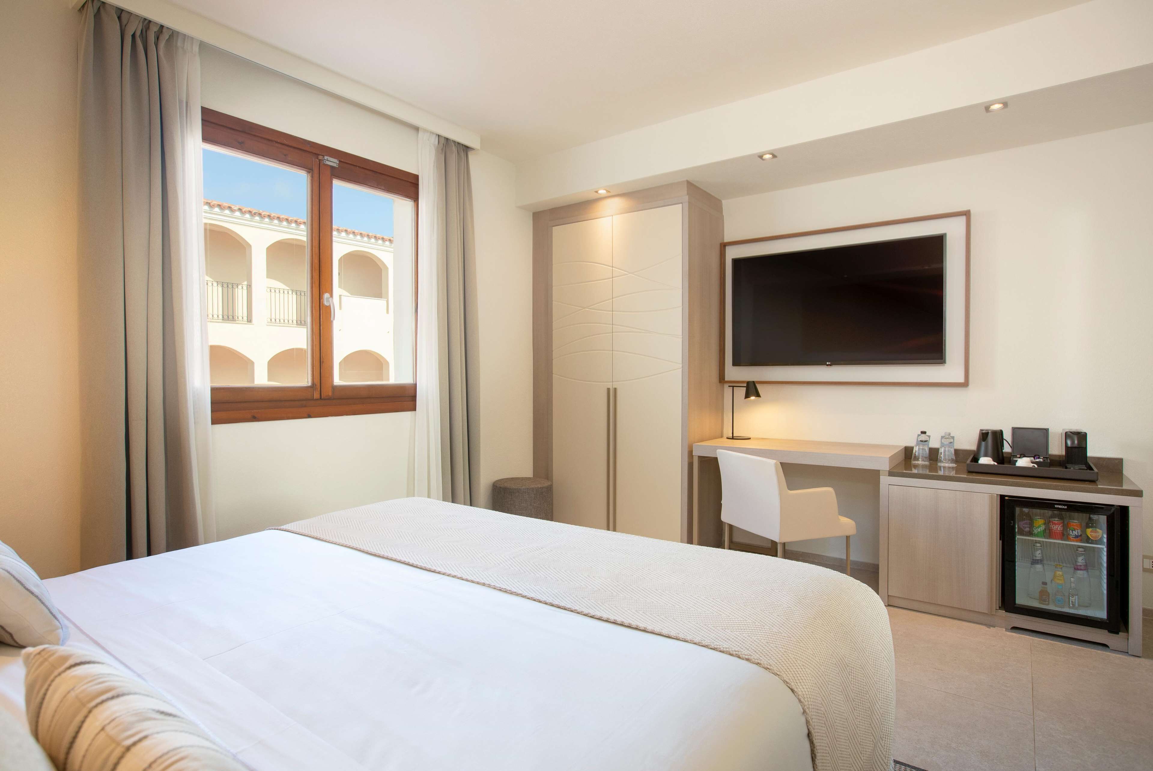 Ett hotellrum med en säng, ett skrivbord, en TV, ett minikylskåp och utsikt över en byggnad med valv.