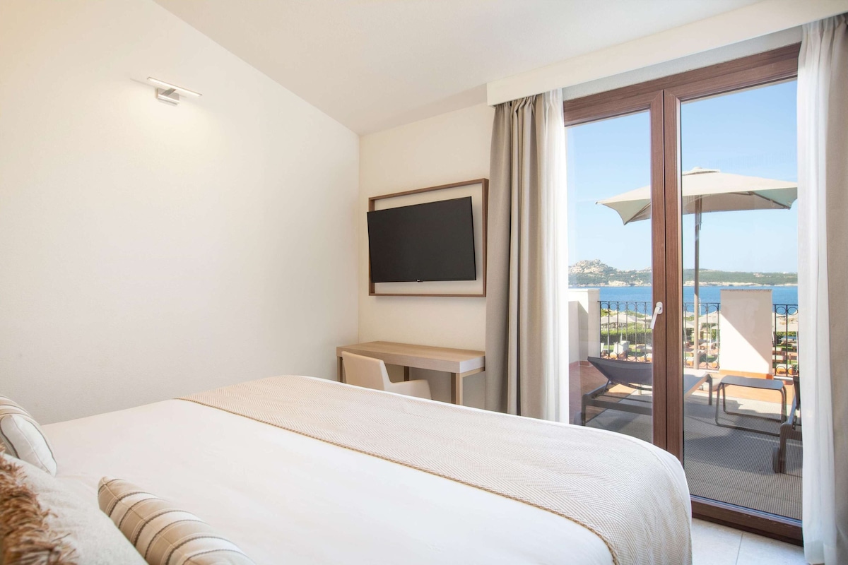 Ein Hotelzimmer mit Bett, Schreibtisch, Stuhl, Fernseher und einem Balkon mit Meerblick.