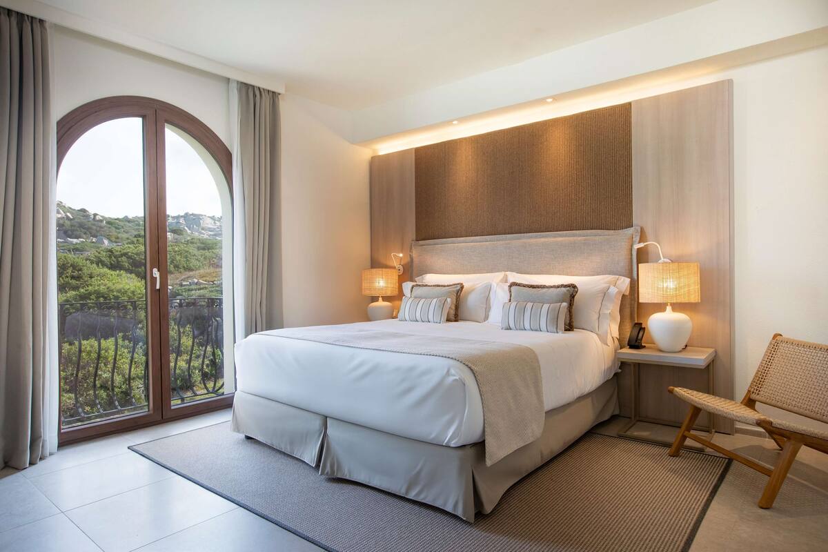 Habitación de hotel moderna con una cama grande, dos lámparas de noche, una silla y vistas a zonas verdes a través de una puerta corrediza.