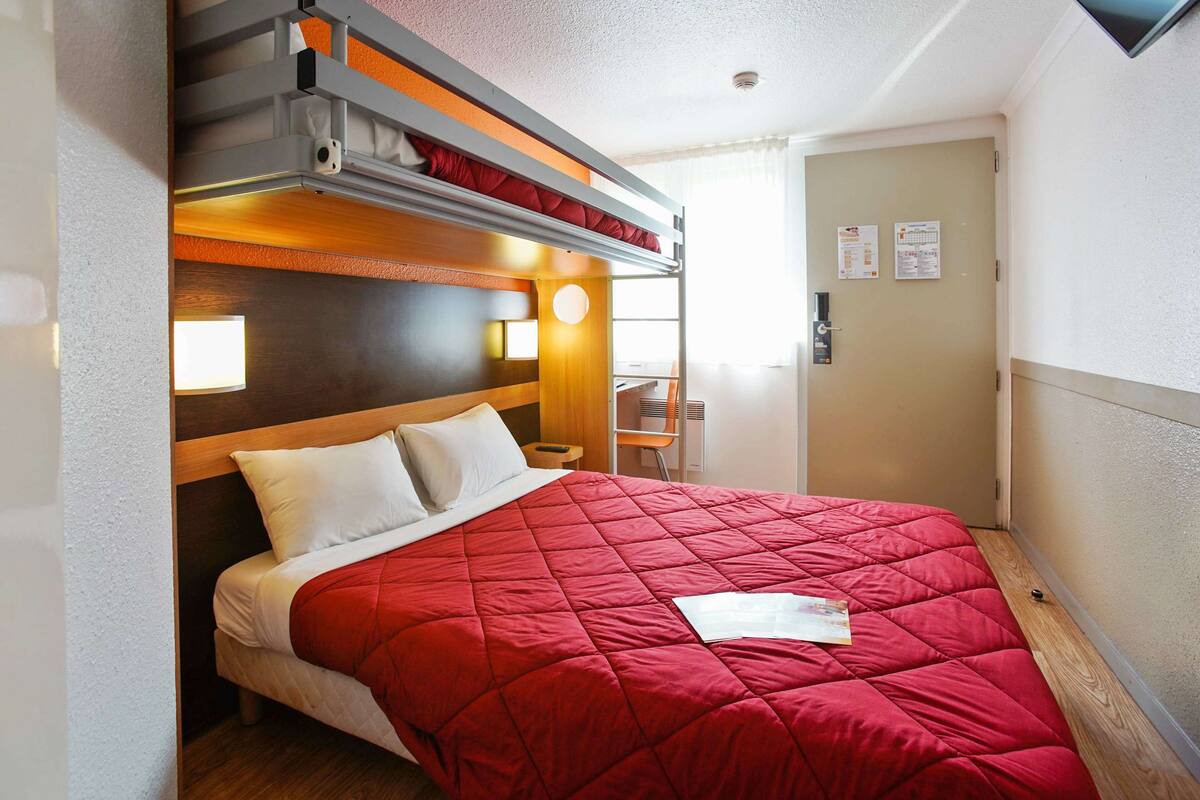 Une chambre d’hôtel compacte avec un lit superposé, une couvre-lit rouge, une tête de lit en bois, une table de chevet avec une lampe et un petit bureau avec une chaise.