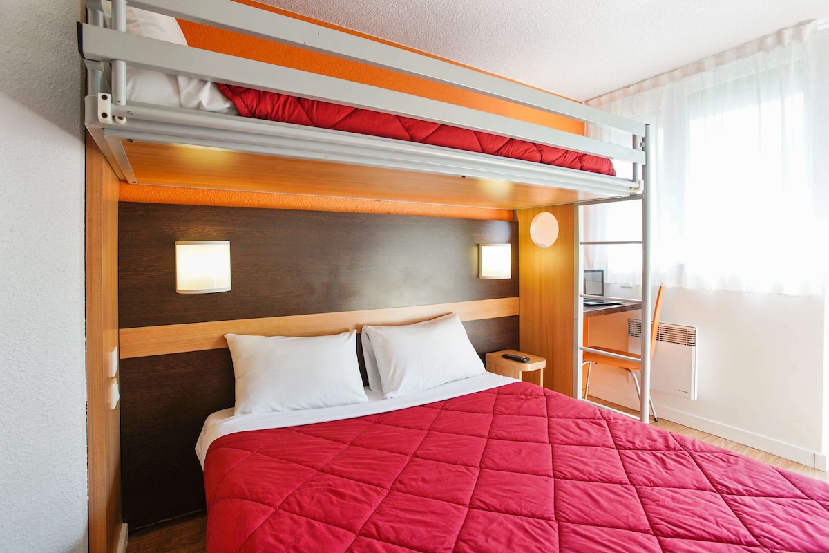 Een hotelkamer met een stapelbed, een rood geborduurde spreidekker, een houten hoofdeinde, twee bedlampjes, een klein tafeltje met een stoel en een raam met gordijnen.